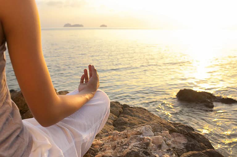 Une personne assise sur un terrain rocheux au bord de l’océan au lever ou au coucher du soleil pratique Dhyana. Ils sont en position jambes croisées, portent un pantalon de couleur claire et un haut sans manches. Leur main repose sur leur genou, les doigts en position mudra. Le soleil projette une lueur dorée sur l'eau, offrant ses bienfaits.