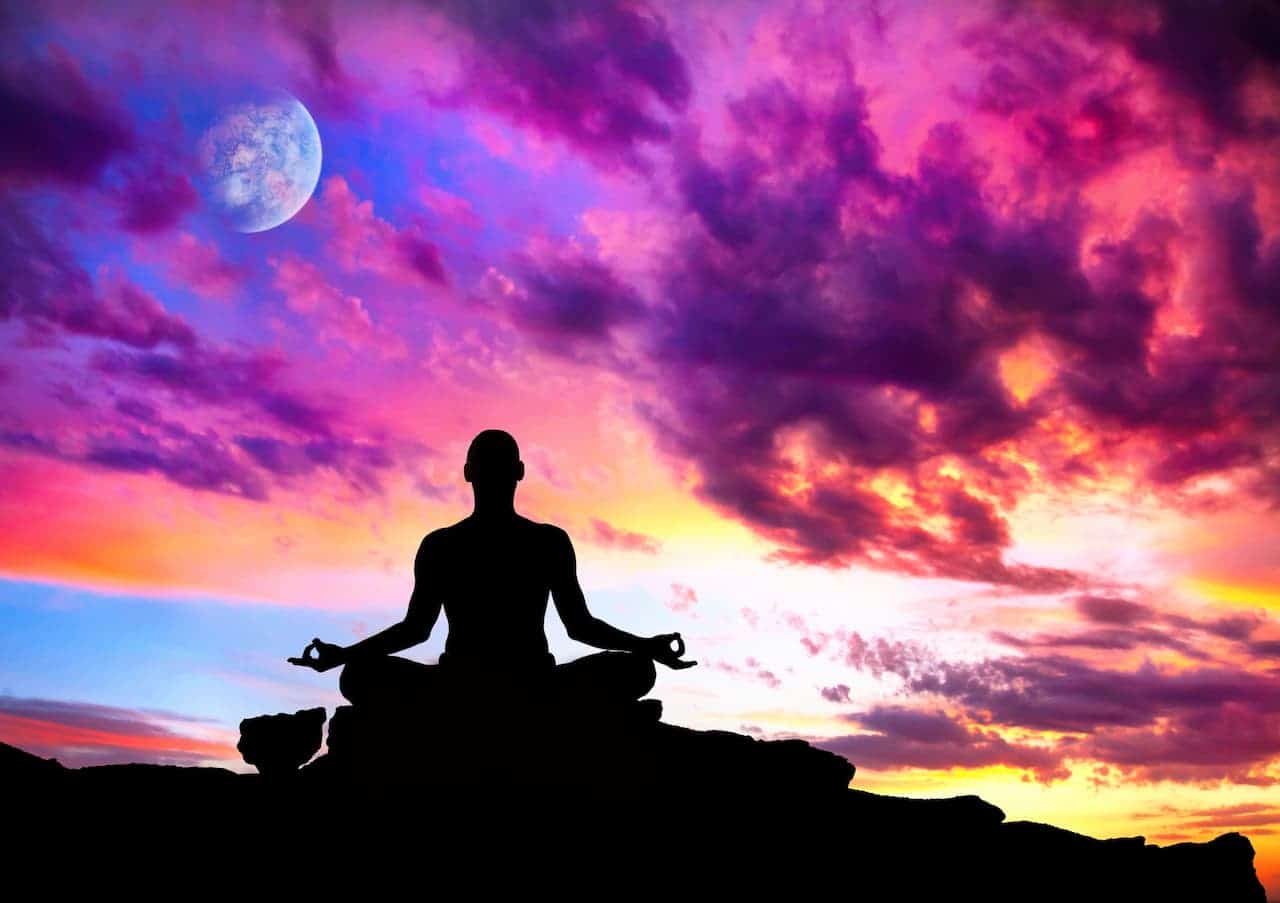 La silhouette d'une personne assise les jambes croisées dans une pose méditative, pratiquant l'Ishta Yoga sur une surface rocheuse. L’arrière-plan est un ciel vibrant et coloré au coucher du soleil avec des nuances de rose, de violet et d’orange. Une pleine lune est visible dans le coin supérieur gauche, partiellement obscurcie par les nuages, évoquant le mystique Rituel de la Lune.