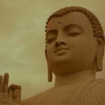 Une vue rapprochée d'une grande statue de Bouddha en pierre contre un ciel nuageux. La statue présente des détails complexes, notamment des cheveux bouclés, des lobes d'oreilles allongés et une expression calme. Une main est levée avec la paume tournée vers l'extérieur, les doigts légèrement fléchis dans un geste transcendant les obstacles intérieurs.