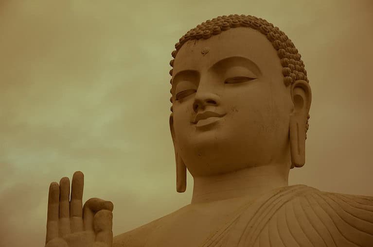 Une vue rapprochée d'une grande statue de Bouddha en pierre contre un ciel nuageux. La statue présente des détails complexes, notamment des cheveux bouclés, des lobes d'oreilles allongés et une expression calme. Une main est levée avec la paume tournée vers l'extérieur, les doigts légèrement fléchis dans un geste transcendant les obstacles intérieurs.