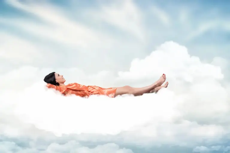 Une femme en robe orange est allongée sur le dos sur un nuage blanc et pelucheux sur fond de ciel bleu parsemé de doux nuages blancs. Elle apparaît détendue, les yeux fermés et les jambes étendues, comme si elle méditait sur «Où Commencent» les limites des rêves. La scène dégage une atmosphère sereine et rêveuse.