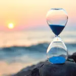 Atteindre la Conscience Kriya Un sablier de sable bleu de la collection « Atteindre la Conscience Kriya » est posé sur un rocher en bord de mer. Le soleil couchant projette des teintes orangées et roses, tandis que de douces vagues en arrière-plan créent une atmosphère sereine.