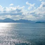 Les Fenêtres vers le Ciel Vue d'un océan serein avec la lumière du soleil se reflétant sur l'eau, vue à travers des fenêtres ouvertes. Les montagnes bordent l'horizon sous un ciel bleu partiellement nuageux. Les cadres des fenêtres sont blancs et la poignée de la fenêtre de droite est visible - vraiment Les Fenêtres vers le Ciel.