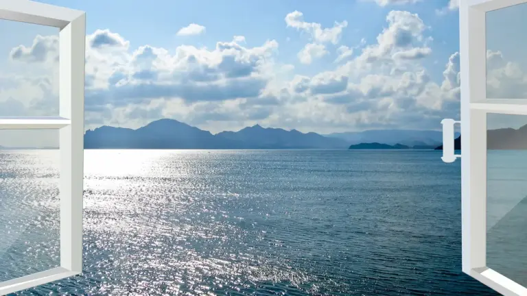 Vue d'un océan serein avec la lumière du soleil se reflétant sur l'eau, vue à travers des fenêtres ouvertes. Les montagnes bordent l'horizon sous un ciel bleu partiellement nuageux. Les cadres des fenêtres sont blancs et la poignée de la fenêtre de droite est visible - vraiment Les Fenêtres vers le Ciel.