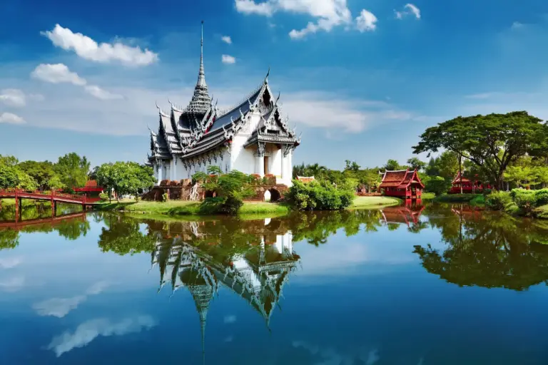 Un temple thaïlandais traditionnel aux toits pointus et ornés se trouve au bord d'un lac calme dans le Royaume Mental, entouré d'une végétation luxuriante. Le temple et les arbres se reflètent clairement dans l'eau, sous un ciel bleu vif parsemé de nuages.