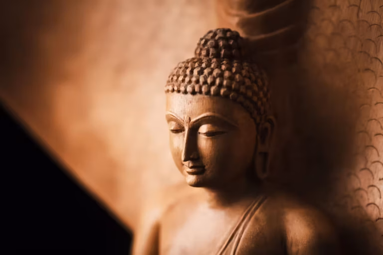 Gros plan sur une statue de Bouddha sereine, aux tons chauds de brun doré. Les yeux fermés et le regard serein, elle incarne le karma et la contemplation paisible. L'arrière-plan légèrement flou met en valeur le travail artisanal minutieux de la statue.