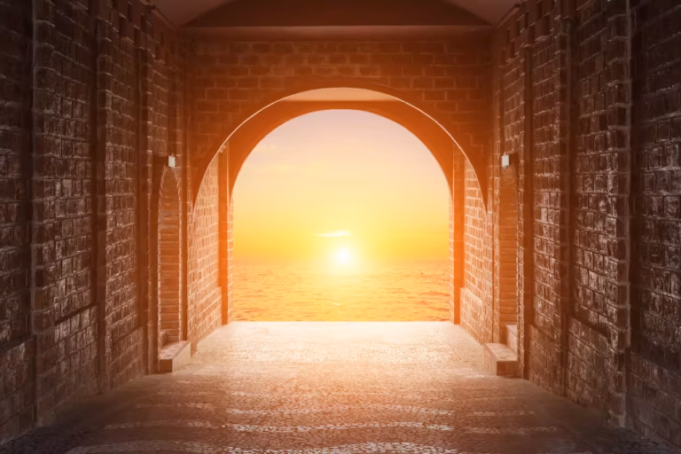 Un couloir en pierre baigné de soleil, doté d’une porte voûtée, s’ouvre sur un magnifique coucher de soleil sur une mer calme, projetant une lumière chaude aux teintes orange et jaune sur les murs en briques et le sol texturé, créant ainsi une atmosphère sereine et accueillante qui invite à une réflexion tranquille sur la destinée humaine.